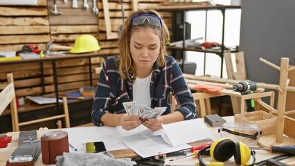 Canvas Print - Serious, young and beautiful hispanic woman carpenter counting heap of dollars on smartphone at her professional woodworking studio, in midst of lumber and furniture. attentive joiner at work.