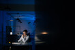 © dikushin - Remote view of serious young business man remote working overtime, learning online late at night in in dark room with neon light using desktop computer at workplace. Concept overwork and deadline.