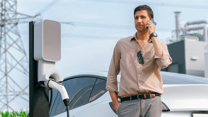 Man talking on the phone while recharge EV car battery at charging station connected to power grid tower electrical as electrical industry for eco friendly car utilization. Panorama Expedient