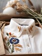 The white linen shirt with handmade embroidery in a giftbox lies on a wooden natural table. Beige embroidered spikelets, cornflowers. Dry herbs, flowers. Stylish modern authentic unique art. Business.