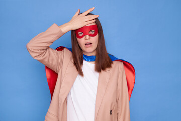 Wall Mural - Displeased disappointed woman wearing superhero costume standing isolated over blue background having troubles showing facepalm gesture forgot something pleasant