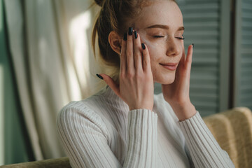 Wall Mural - young happy woman applying cosmetic face cream on skin relaxing on sofa at home in living room. skin