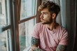 Calm man in pink t-shirt with tattoo on arms looking through window