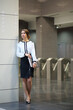 The business woman in white blouse and black skirt with the folder standing in the lobby in front of turnstiles in business center