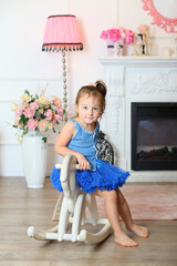 Little girl sitting on rocking horse in blue skirt in front of fireplace in the living room in romantic style