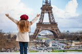Fototapeta Paryż - A happy tourist girl in a trench coat and with a red beret hat looks at the famous Eiffel Tower in Paris, France