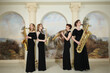 Four women in black dresses play saxophones in studio with pilasters