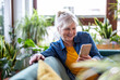 © pikselstock - Smiling senior woman using smart phone while sitting on sofa at home