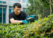 product photography of a man using powerful and imposing garden power tool, a cordless hedge trimmer with a long blade to cut the top of green hedges in a mid size modern house garden