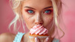 Sugary sprinkles on a woman's face highlight her blue eyes, blonde hair as she enjoys a pink frosted cupcake against a pink background.