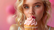 A woman with striking blue eyes and blonde hair savours a pink frosted cupcake against a pink backdrop.