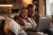 © zephyr_moonstone - Photo of a happy middle-aged couple sitting on the sofa in a modern living room and using a laptop