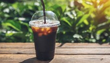 Close Up Of Take Away Plastic Cup Of Iced Black Coffee Americano On Wooden Table With Garden Green Nature Background