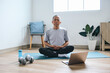 Asian senior man meditating on mat, learning yoga online class using laptop at home