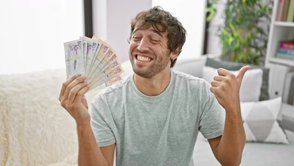 Poster - Cheerful young man at home, confidently holding colombian pesos, pointing thumb up with a beaming smile and open mouth gesture