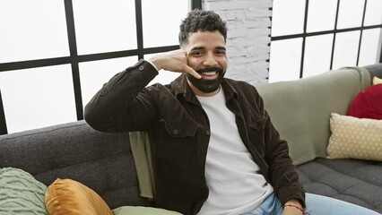 Sticker - Cheerful young man in his indoor apartment playfully making talking gestures with his fingers as a phone, communicating joyfully and confidently at home.