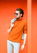 Stylish young man in an orange sweater and summer suit and sunglasses against the background of an orange wall on a summer day.