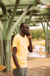 A dark-skinned man in a yellow T-shirt talks on the phone on a summer day in the park.