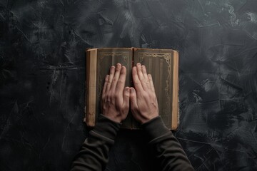 Wall Mural - Religious man praying with separated hands up and religious book in the background on black table. Top view.