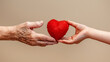 A plush red heart is exchanged between a wrinkled elderly hand and a smooth young hand.