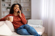 Cheerful black woman enjoying mobile phone conversation and drinking coffee at home