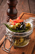 Close-up image showcasing a mix of green and black olives, red pepper slices, and herbs marinated together in a sealed glass jar