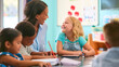 Female Primary Or Elementary School Teacher Helping Students At Desk In Multi-Cultural Class 