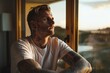 calm man in a casual t-shirt, arms adorned with detailed tattoos, gazing through a large window at a tranquil morning landscape, reflective atmosphere.