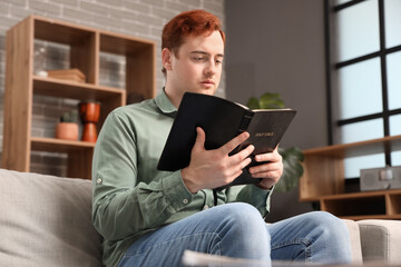 Wall Mural - Young man with Holy Bible praying on sofa at home