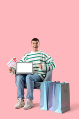 Poster - Young man with gift card, laptop and shopping bags sitting in armchair on pink background