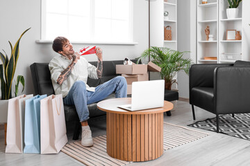 Poster - Young tattooed man with gift voucher and shopping bags at home