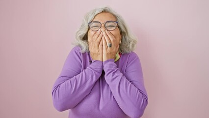 Canvas Print - Cheerful mature woman with grey hair, standing while wearing glasses and playfully covering her mouth, caught laughing amidst juicy gossip over pink isolated background