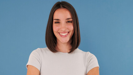 Wall Mural - Close up, beautiful brunette young woman dressed in white top looking at camera and smiling widely isolated on blue background in studio