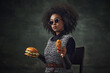 Fast food ads. African woman, medieval warrior in chainmail, sunglasses, holding burger and chicken nuggets over dark vintage background. Concept of comparison of eras, history, creative art, remake