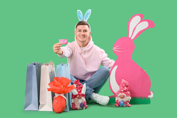 Wall Mural - Young man in bunny ears with credit card, shopping bags and decor on green background. Easter Sale