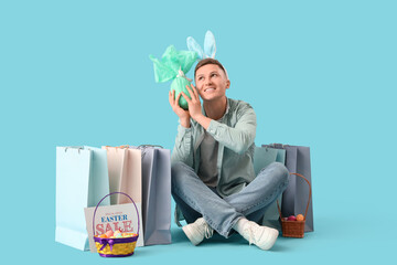 Sticker - Young man in bunny ears with gift egg and shopping bags sitting on blue background. Easter Sale