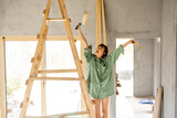 Fototapeta Paryż - Portrait of a young cute woman standing happily on a ladder with paint roller during repairing process of a house. Creative process of home renovation and repair concept