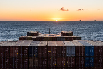 View on the containers loaded on deck of the large cargo ship. She is sailing through calm, blue ocean toward the sunset.