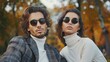 Stylish man and woman in sunglasses pose for the camera outdoors among trees in autumn colors. A handsome male model is dressed elegantly and his beautiful girlfriend is wearing a white jumper.