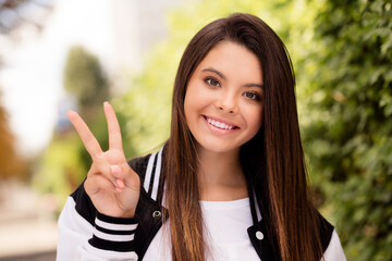 Wall Mural - Portrait of good mood adorable girl with brunette hairstyle wear school uniform showing v-sign symbol say hello on city street outdoors