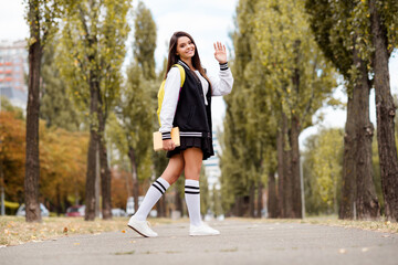 Poster - Full length photo of polite girl wear school uniform long socks rucksack go away with book waving you palm on green city street outdoors