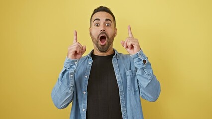 Poster - Young, joyful hispanic male in denim shirt, expressing amazement and cheer, looking up and pointing, stood isolated on sunlit yellow wall background.