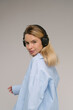 Listening to music. Young Caucasian blonde woman looks at the camera half a turn from  with flirting smile with headphones on her neck. Blue casual shirt. Grey background studio shot music audio theme