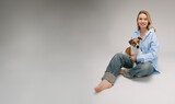 Fototapeta Konie - Smiling blonde woman in a blue shirt sits on the floor, the dog sitting on her laps. Funny cute friends studio portrait. Grey background. Large size banner