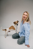 Fototapeta Konie - laughing blonde woman in a blue shirt sits on the floor, the dog stands with its paws on her leg. Funny cute friends studio portrait. Grey background. 