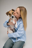 Fototapeta Konie - Kissing a pet. Blonde beautiful woman in blue shirt holding her dog Jack Russell terrier and kissing. Studio shot. Grey background