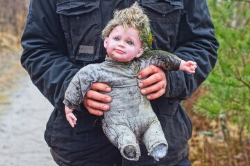 Wall Mural - old dirty torn children's toy small in gray clothes with a plastic face and hair doll of a girl in the hands of a man dressed in a black jacket on the street during the day