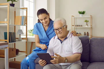 Wall Mural - Young pretty brunette nurse at home communicates with an elderly man with glasses, showing him something on a tablet, conveying good mood and positive attitude. The concept of psychological health.