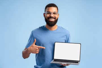 Smiling African American man wearing eyeglasses holding laptop pointing finger at white blank screen