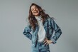 A happy woman in jeans and an oversized denim jacket stands against the background of gray